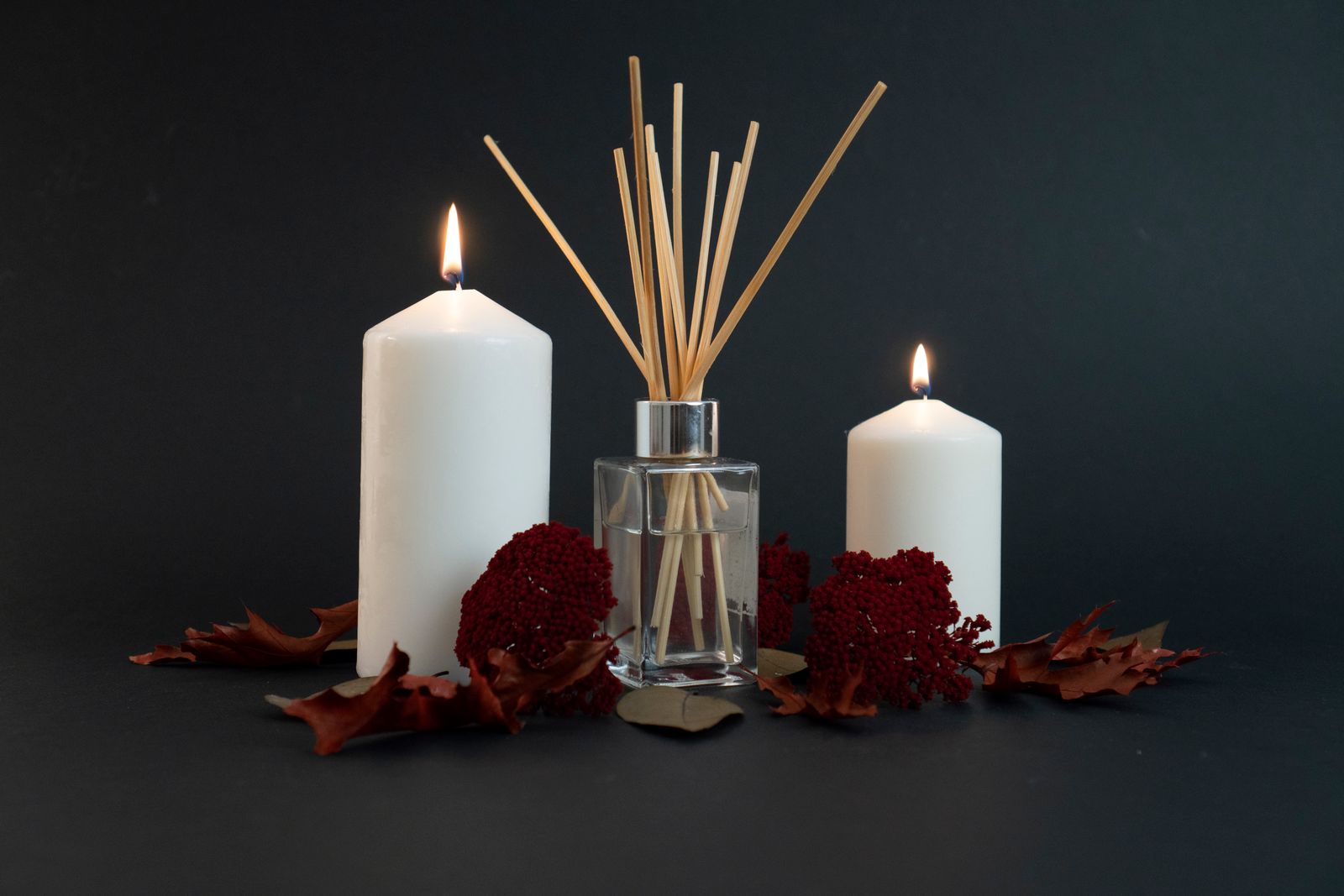 A glass reed diffuser between two lit white pillar candles with dried red flowers and autumn leaves on a dark background