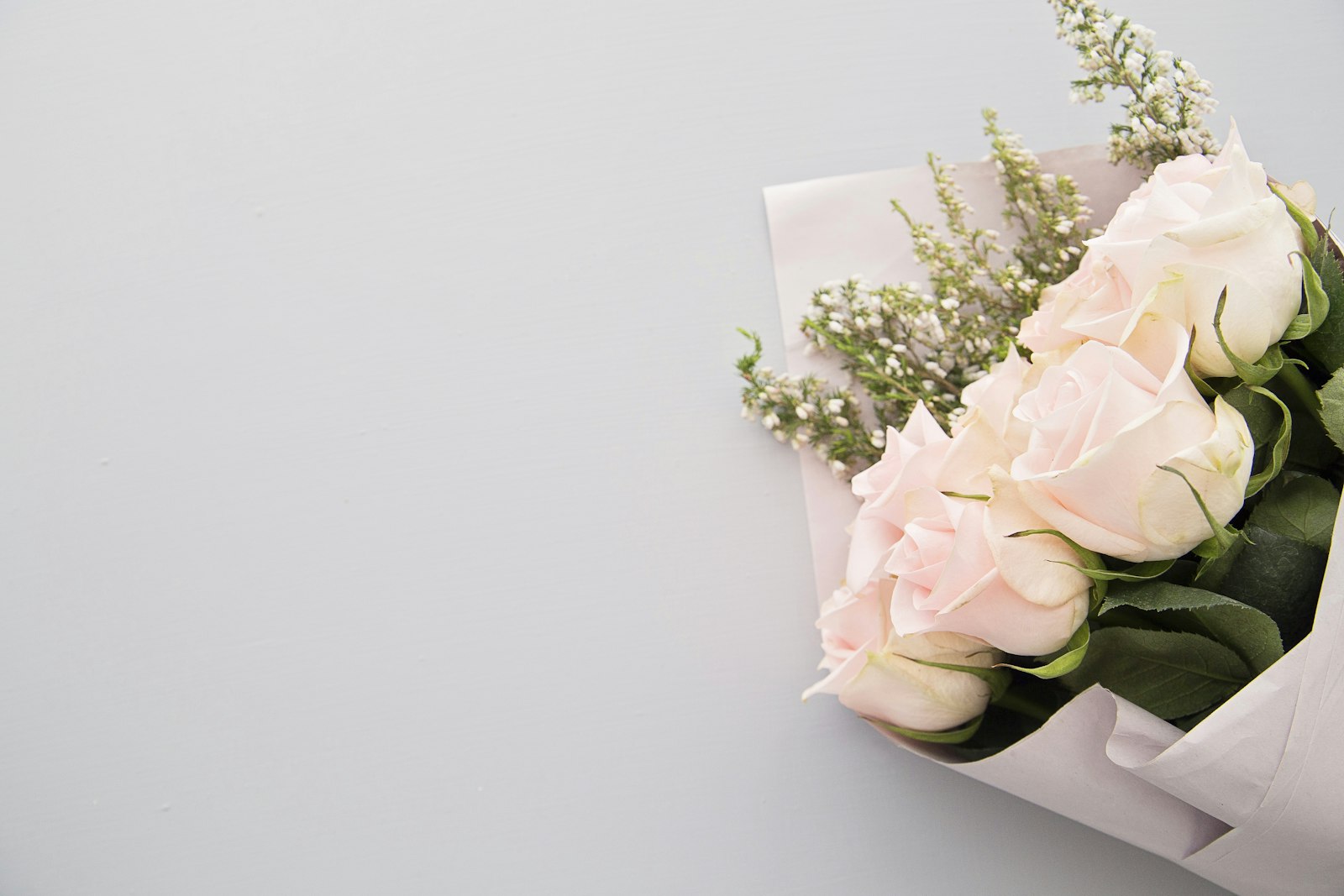 A bouquet of pale pink roses and white filler flowers wrapped in paper on a grey surface