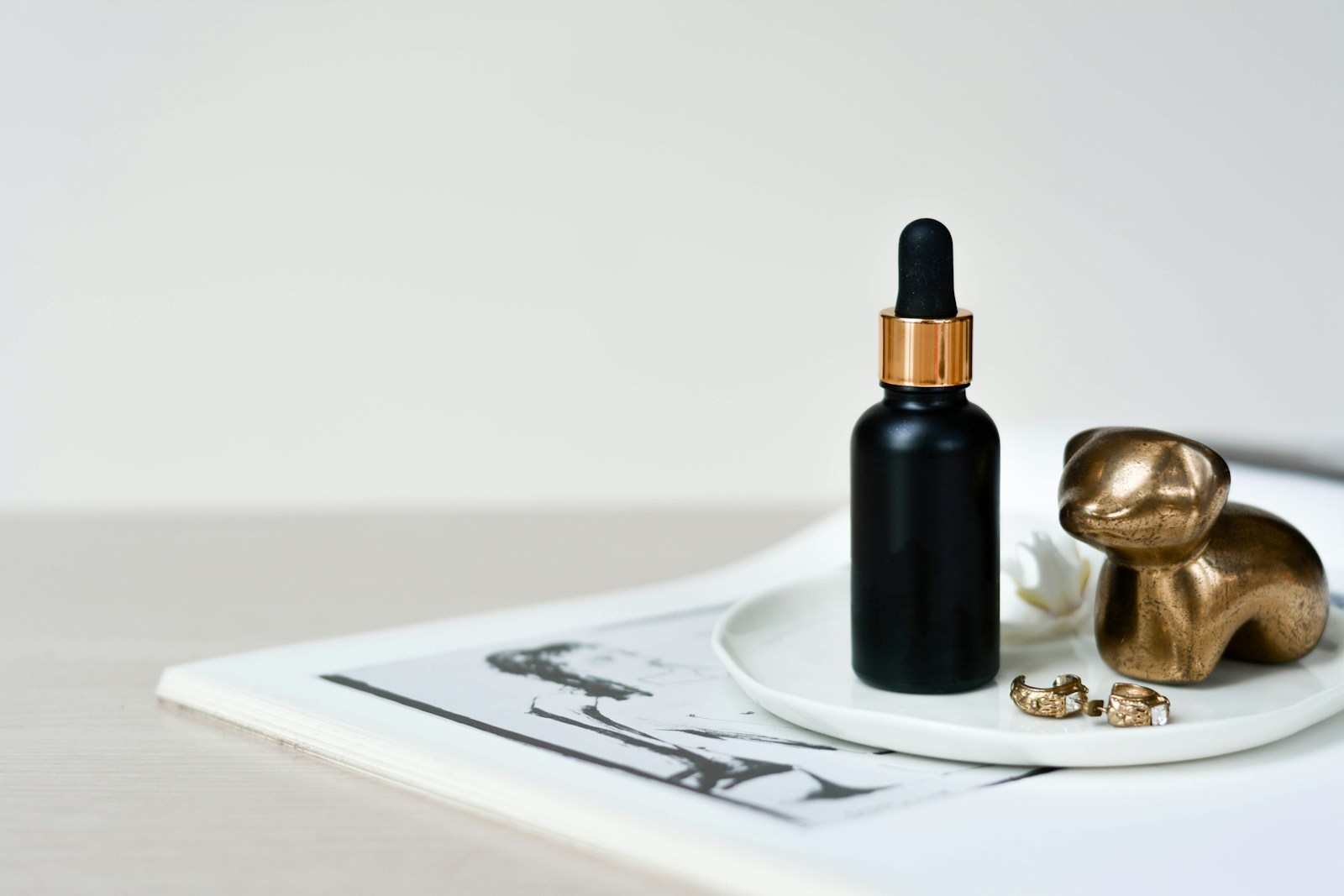 A black dropper bottle of serum on a white dish beside gold earrings