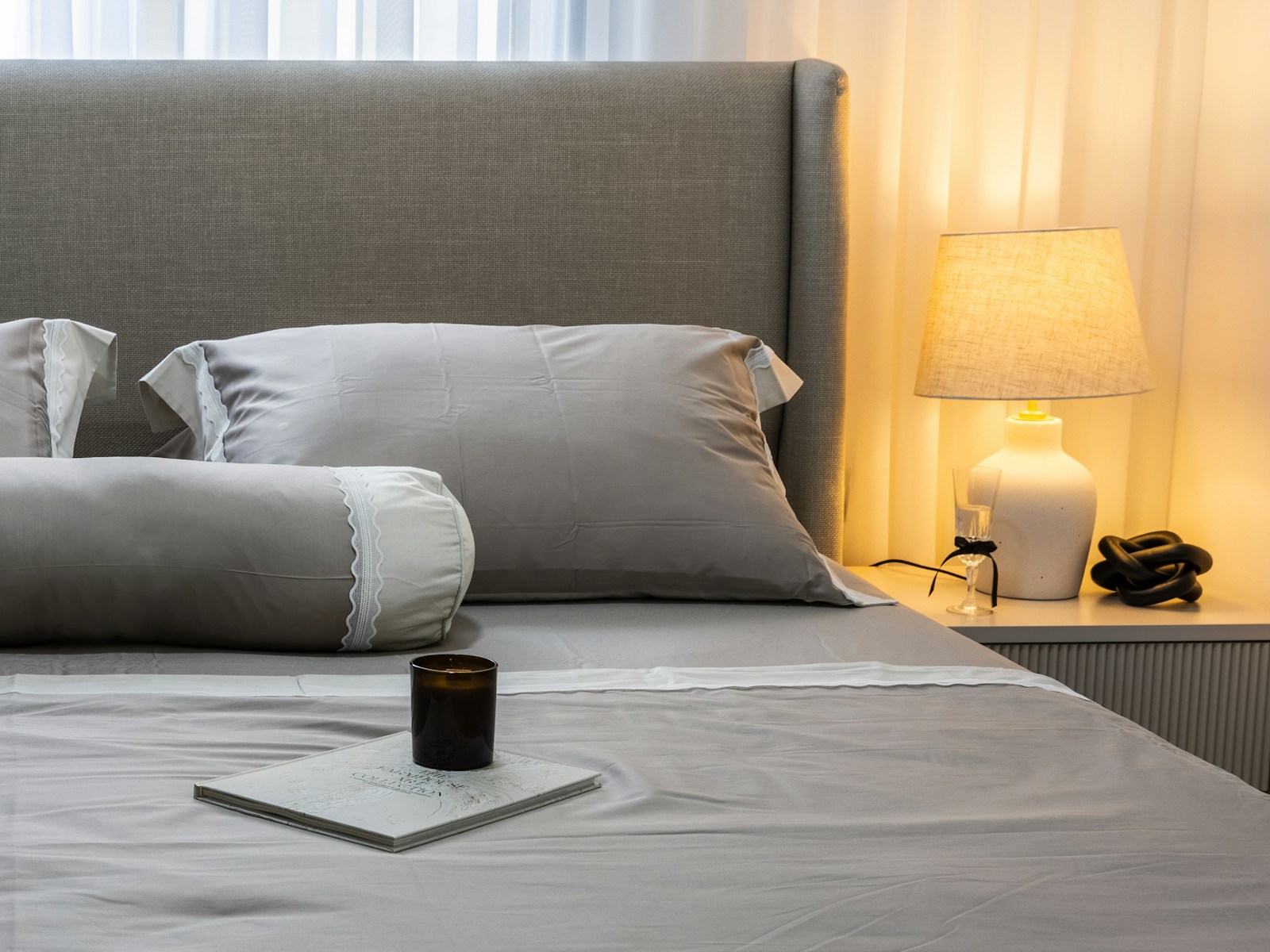 A neatly made bed with grey sheets and pillows beside a lit bedside lamp