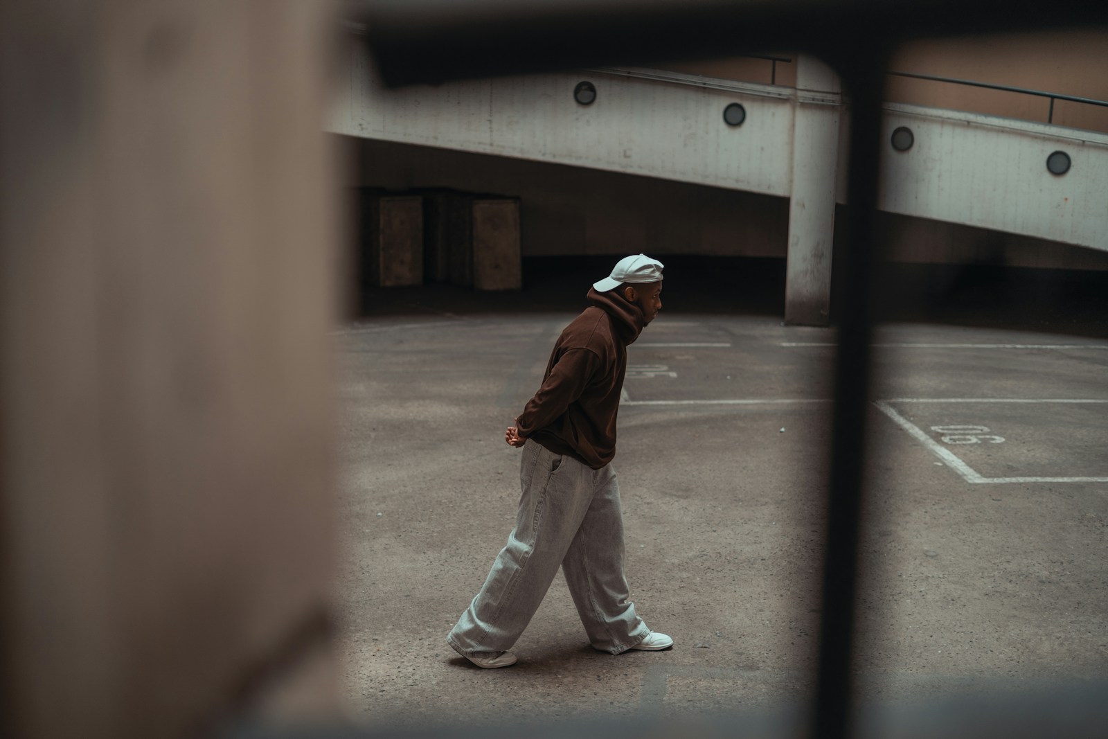 Person in a brown hoodie, cap and grey joggers walking across an empty concrete space