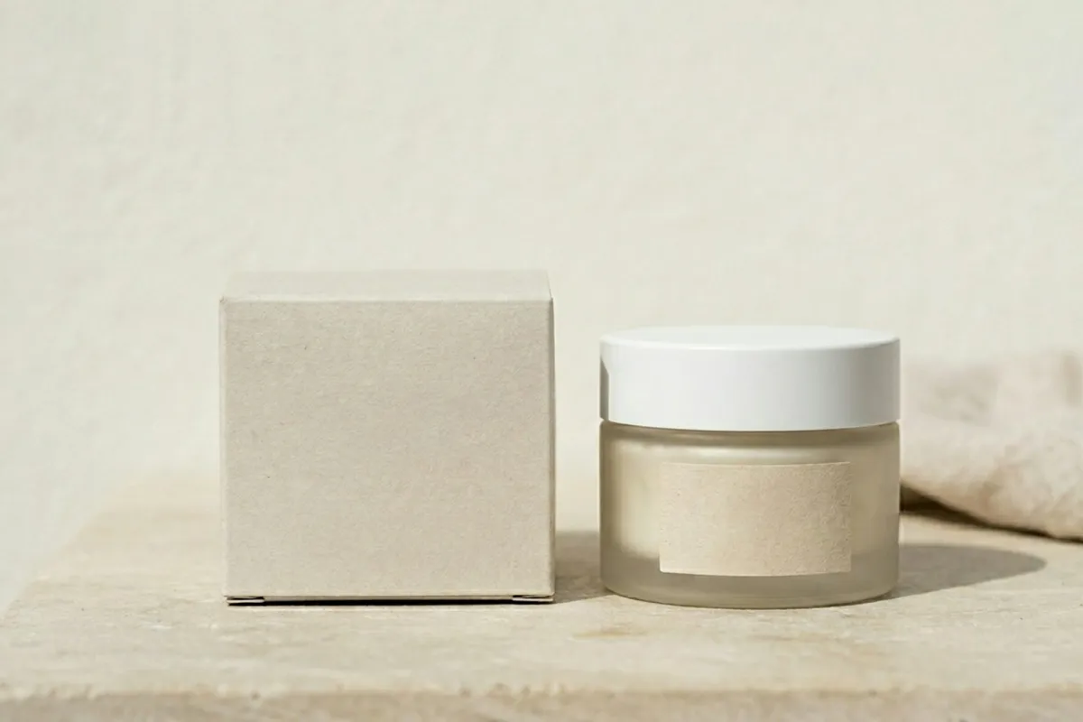 A frosted glass cream jar with a white lid beside a plain cardboard box A frosted glass cream jar with a white lid beside a plain cardboard box