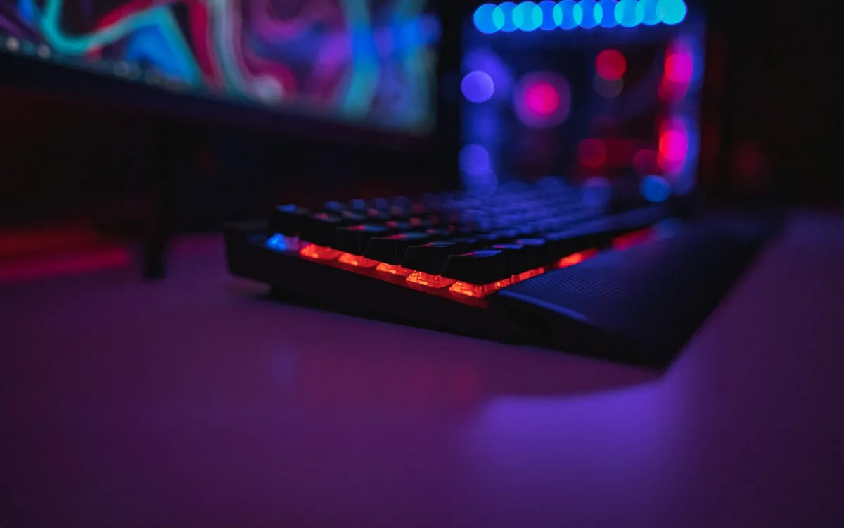 A backlit gaming keyboard on a desk in a dark room lit by coloured lights