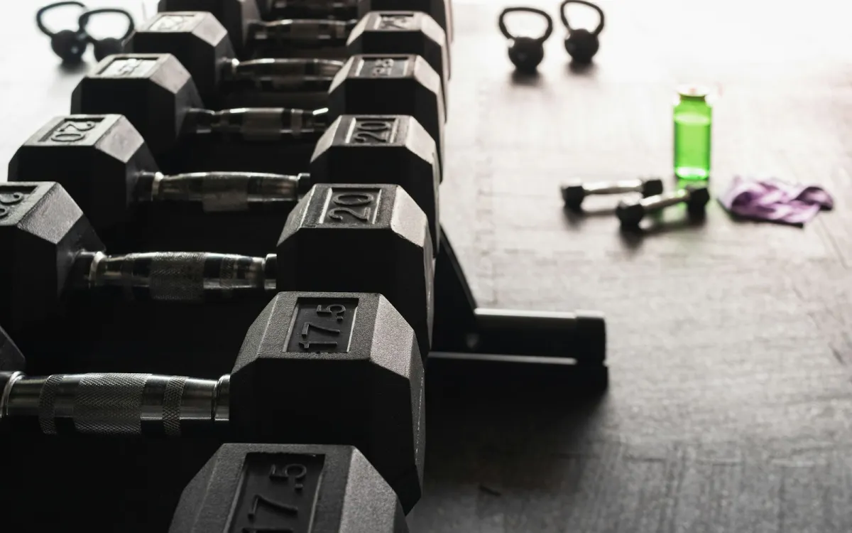 A rack of black hex dumbbells with kettlebells and a water bottle on a gym floor