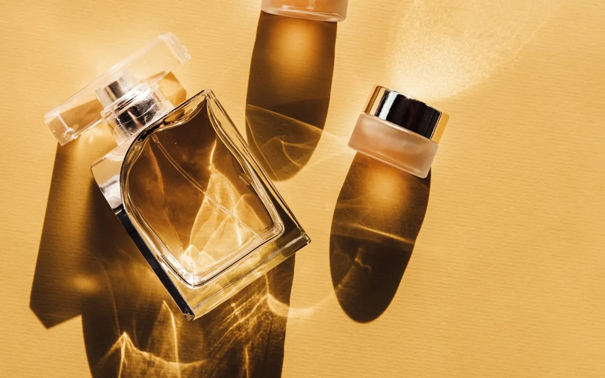 A glass perfume bottle and two small cosmetic jars casting shadows on a golden yellow surface A glass perfume bottle and two small cosmetic jars casting shadows on a golden yellow surface