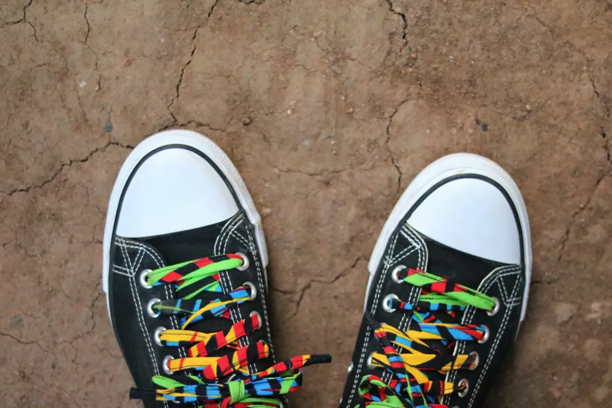 A pair of black canvas trainers with colourful patterned laces on a dusty floor