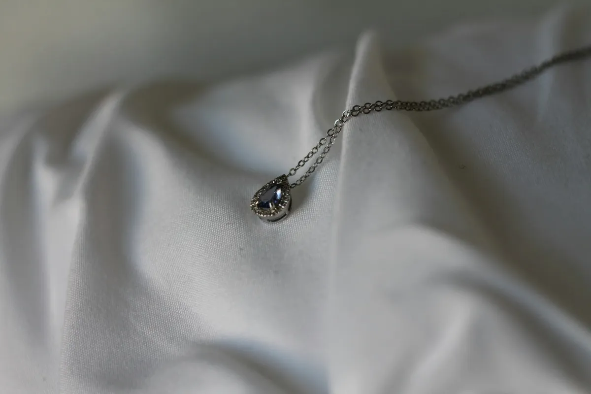 A silver pendant necklace with a small teardrop stone resting on folded white fabric A silver pendant necklace with a small teardrop stone resting on folded white fabric