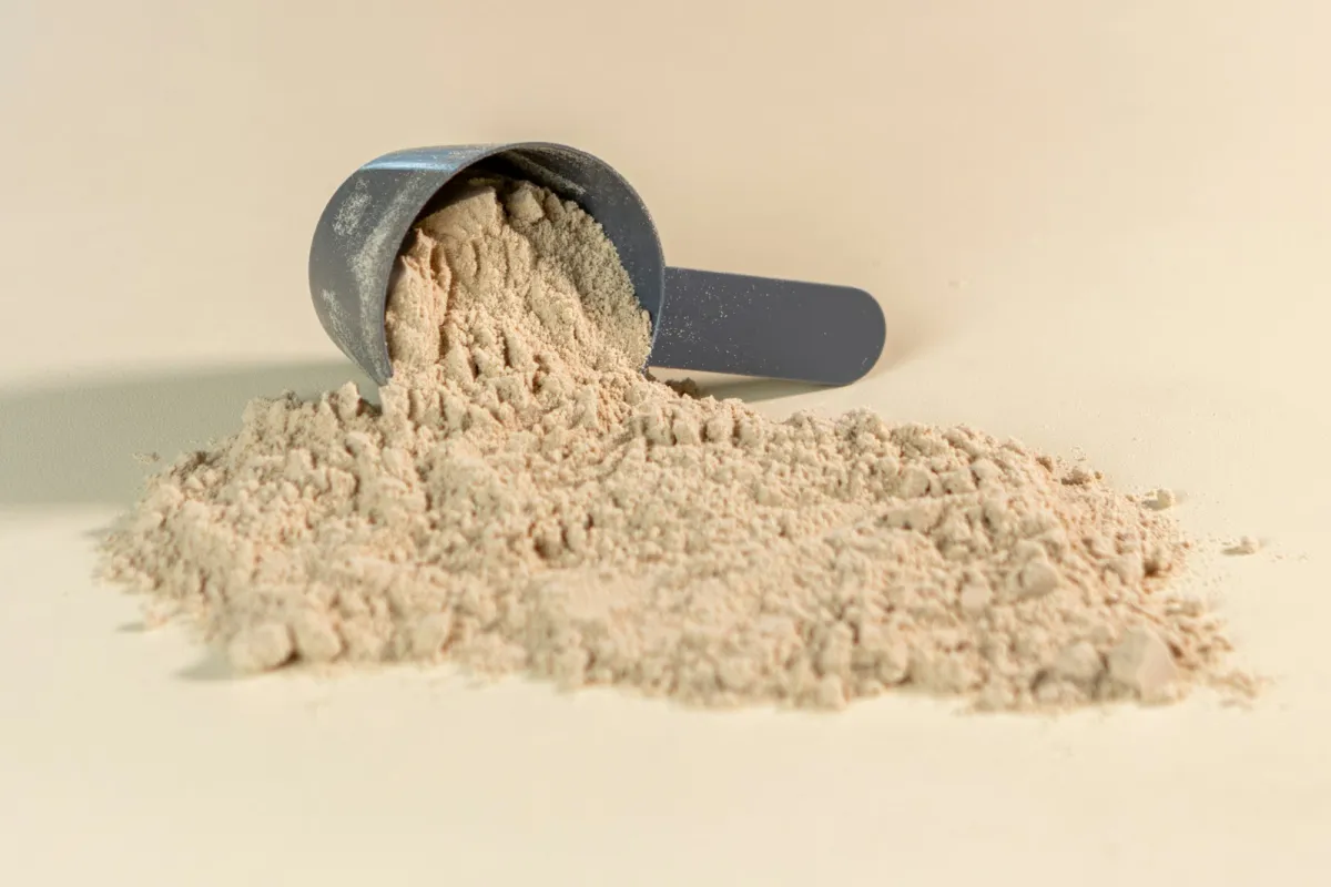 A grey scoop tipping protein powder onto a pale surface A grey scoop tipping protein powder onto a pale surface