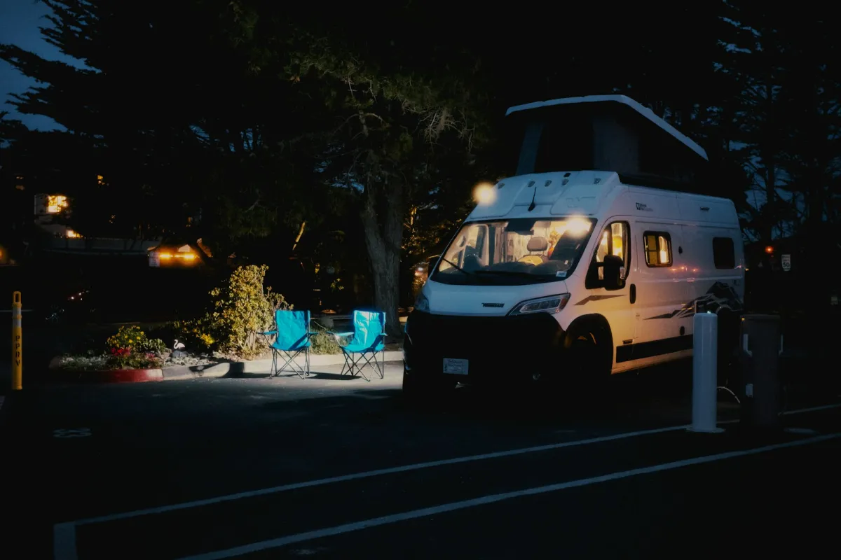 A campervan with its lights on beside two camping chairs at a campsite at night A campervan with its lights on beside two camping chairs at a campsite at night