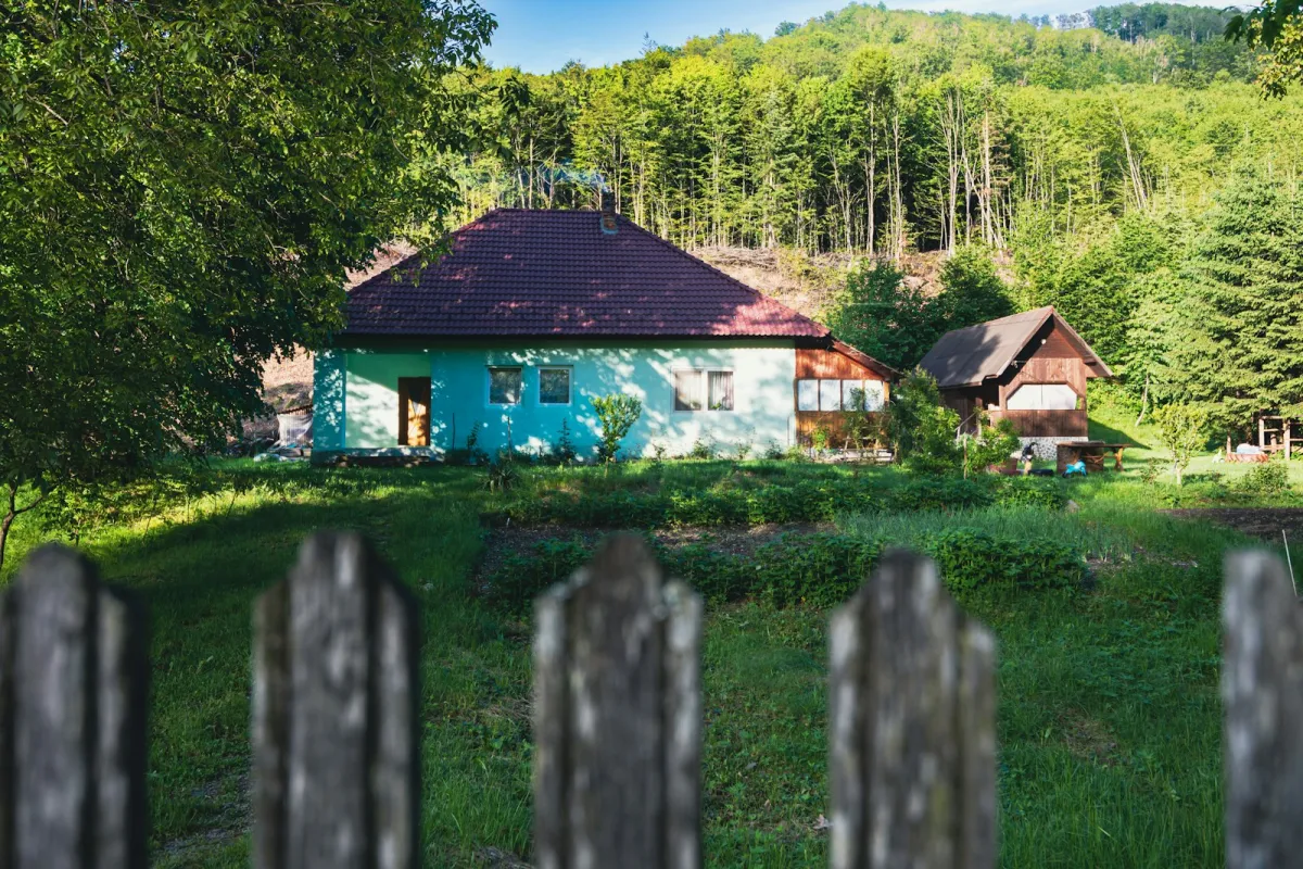 A small house with a red roof in a garden clearing below a wooded hillside