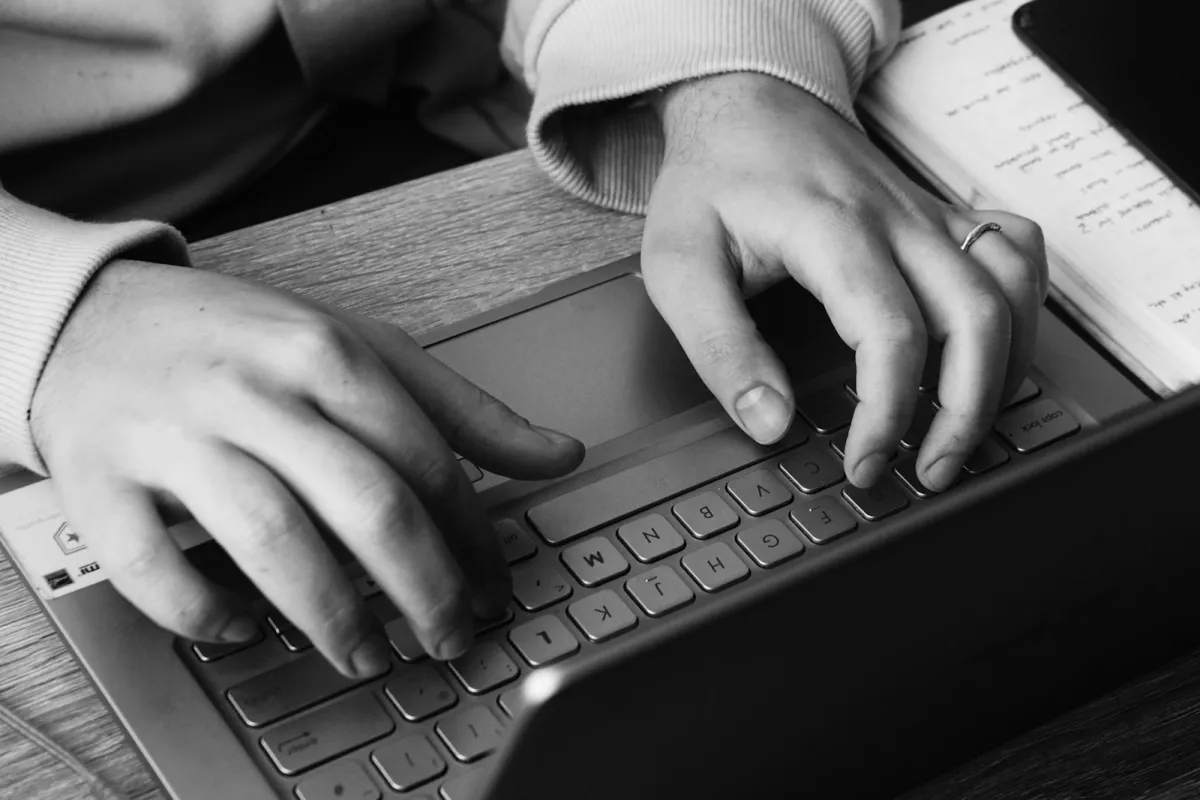 Black and white close-up of hands typing on a laptop beside an open notebook Black and white close-up of hands typing on a laptop beside an open notebook