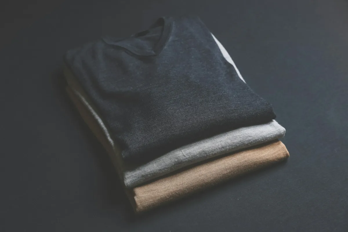 Three folded V-neck jumpers in navy, grey and camel stacked on a dark surface