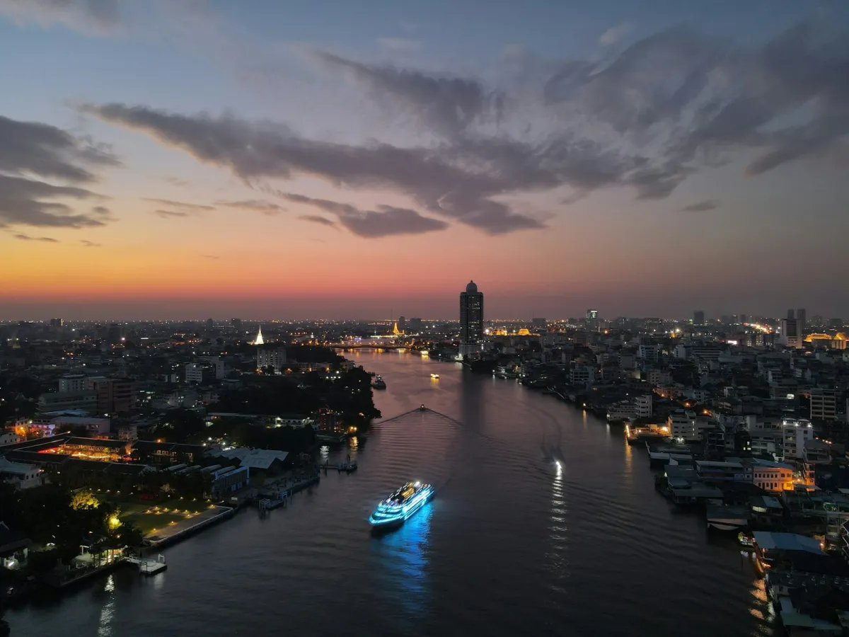 Boats on the Chao Phraya river through Bangkok at dusk, with the city lights coming on