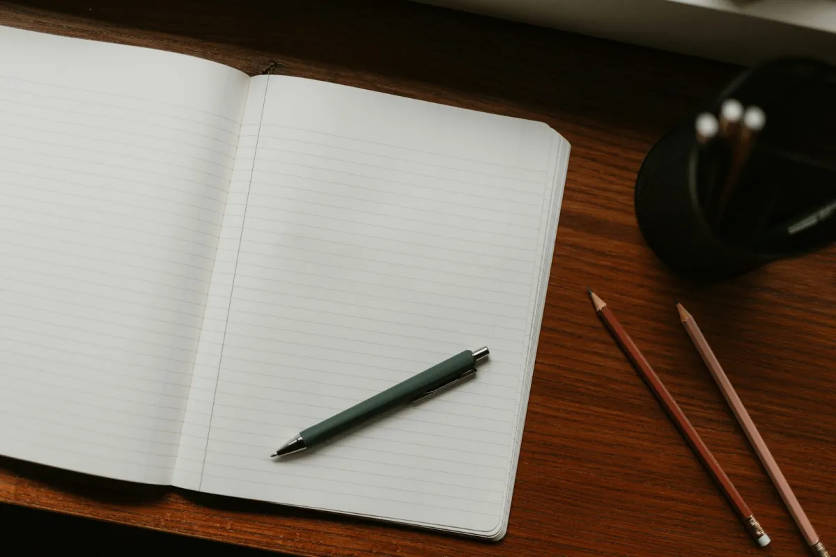 An open lined notebook with a pen on a wooden desk beside two pencils An open lined notebook with a pen on a wooden desk beside two pencils