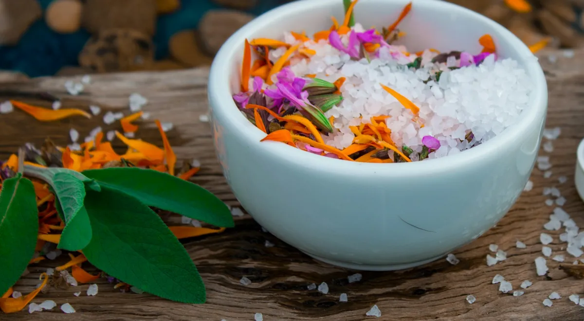 White bowl of coarse bath salts mixed with dried petals on a rustic wooden board