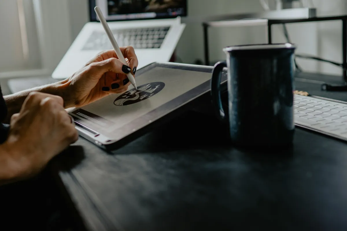 Hands drawing an illustration on a tablet with a stylus at a desk, next to a mug and keyboard
