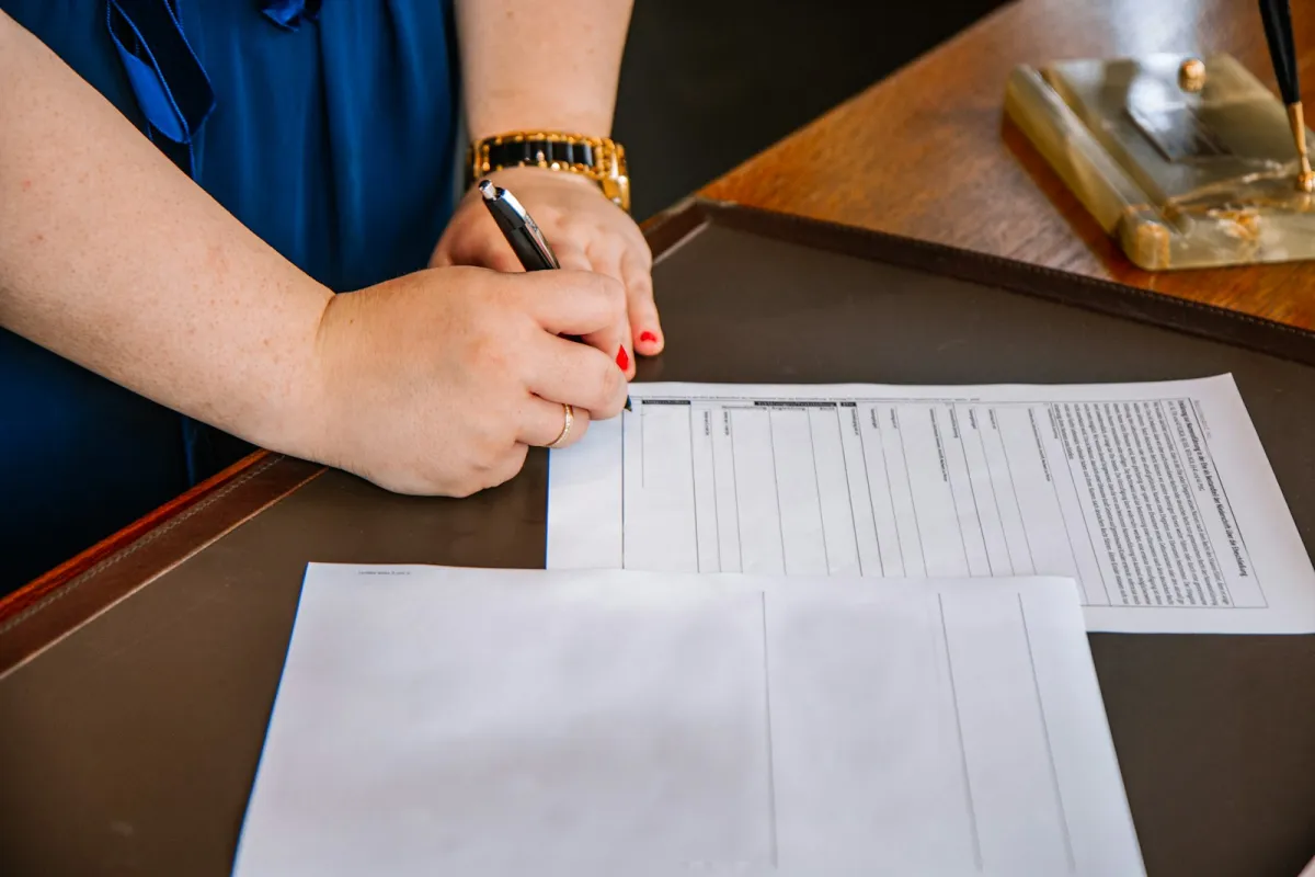 A person's hand signing paperwork with a pen on a desk