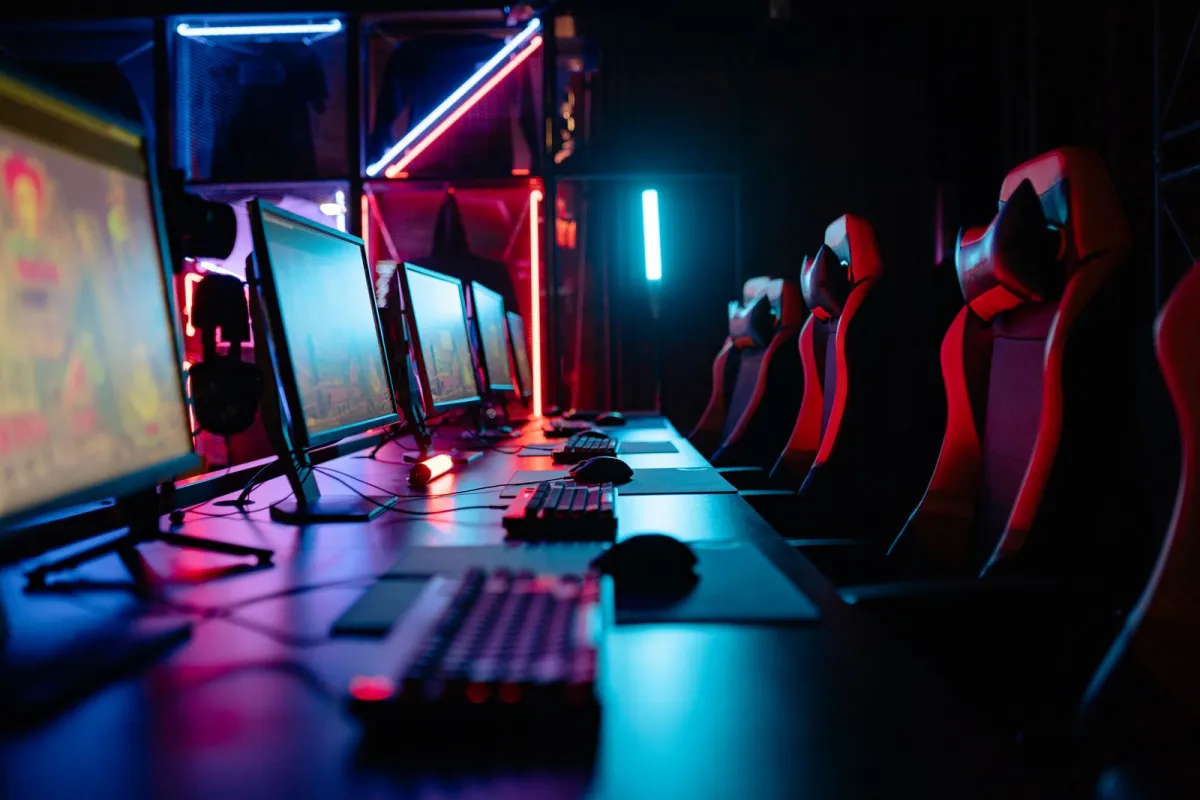 A row of red and black gaming chairs facing computer screens in a dimly lit room with neon lights A row of red and black gaming chairs facing computer screens in a dimly lit room with neon lights