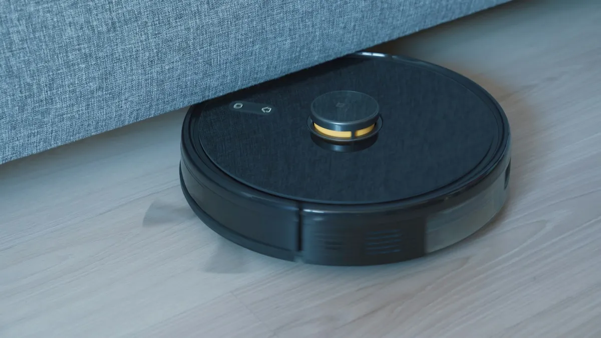 A black robot vacuum cleaning a light wooden floor beside a grey sofa