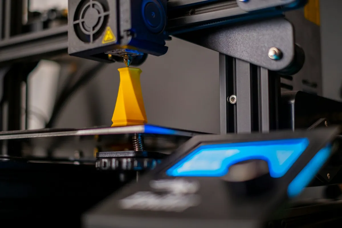 A 3D printer nozzle building a yellow plastic model, lit by a blue display A 3D printer nozzle building a yellow plastic model, lit by a blue display