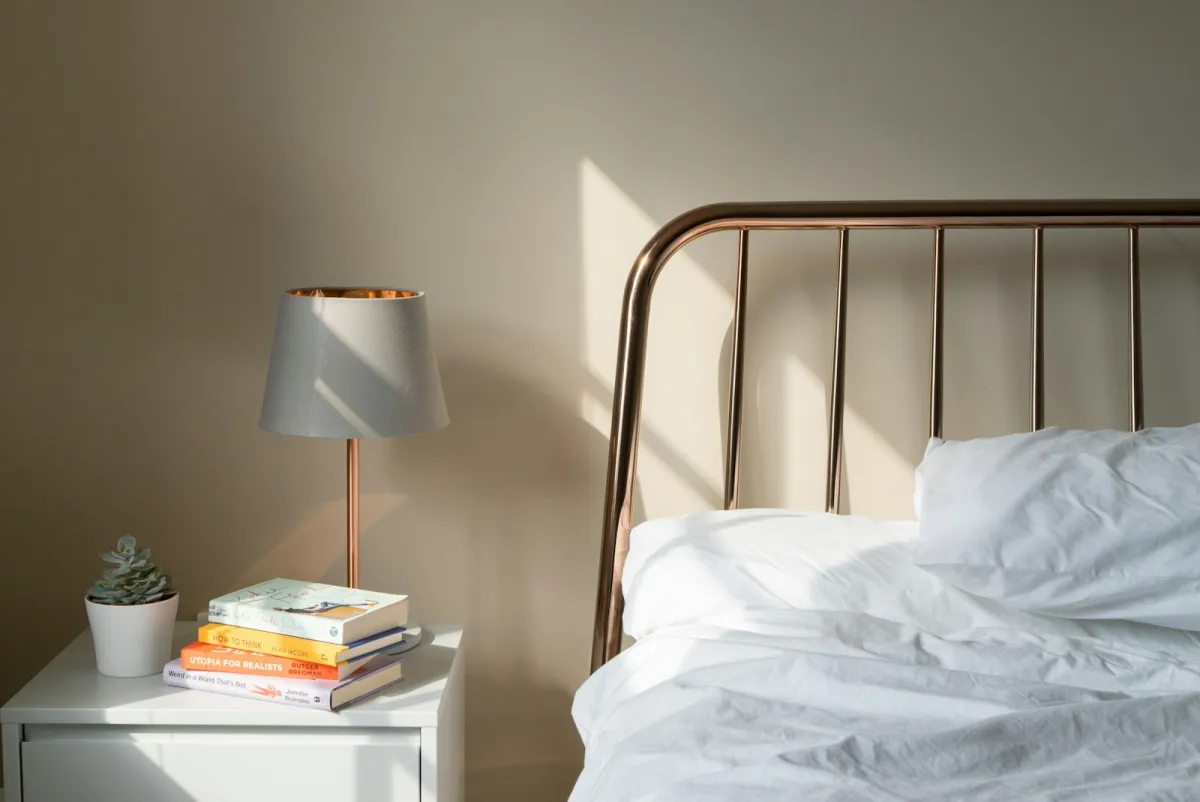 A white bedside table with a lamp and books next to a bed with white linen and a metal headboard A white bedside table with a lamp and books next to a bed with white linen and a metal headboard