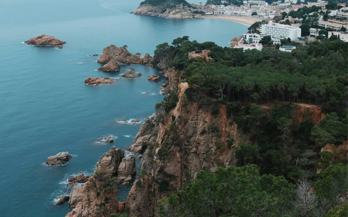 Pine-covered cliffs above the sea on the Costa Brava, with a beach town in the distance Pine-covered cliffs above the sea on the Costa Brava, with a beach town in the distance