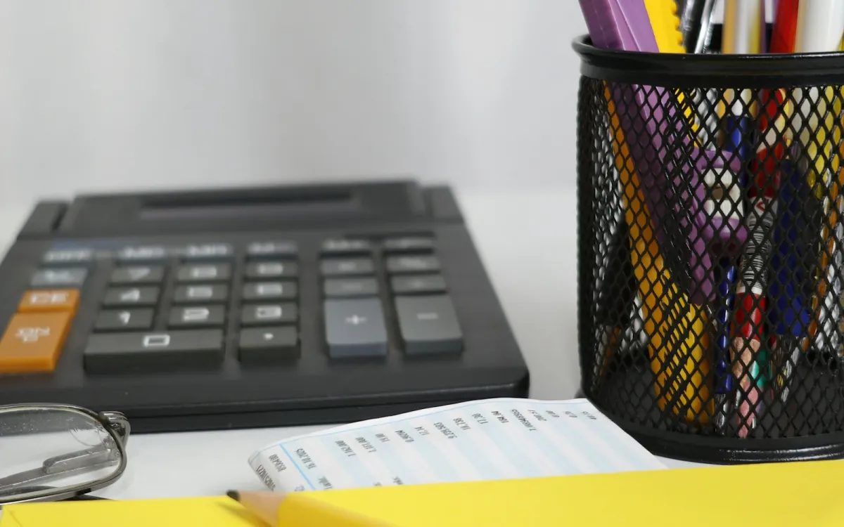 A calculator, a receipt, glasses and a pot of pens on a desk beside a yellow envelope A calculator, a receipt, glasses and a pot of pens on a desk beside a yellow envelope