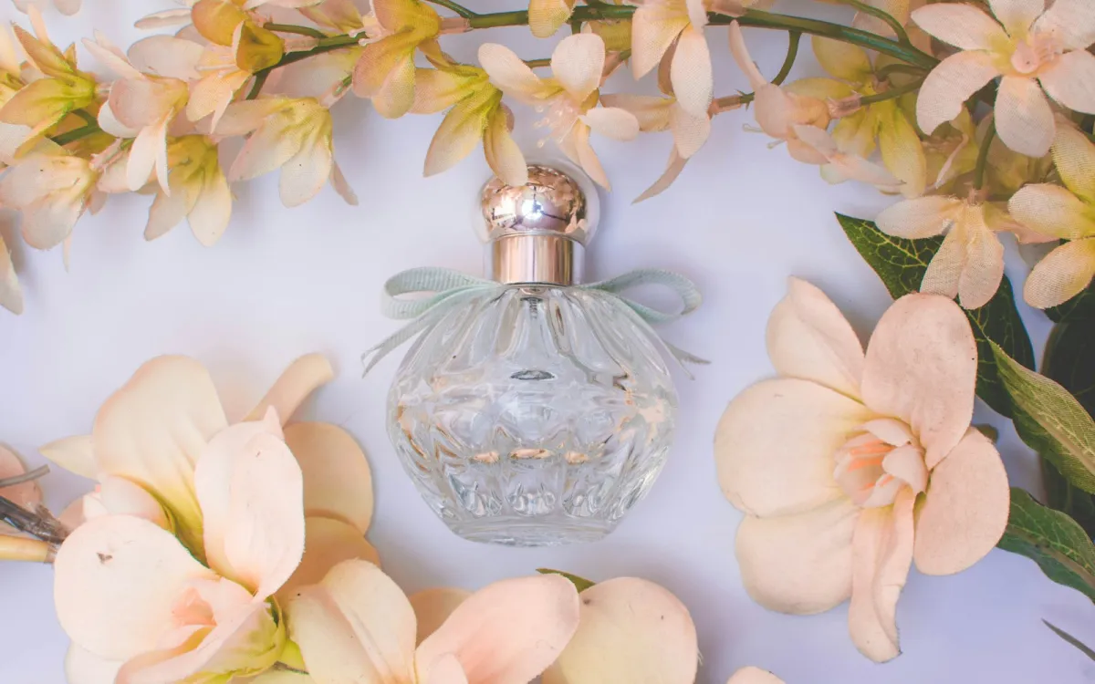 A clear glass perfume bottle with a ribbon, surrounded by pale flowers A clear glass perfume bottle with a ribbon, surrounded by pale flowers