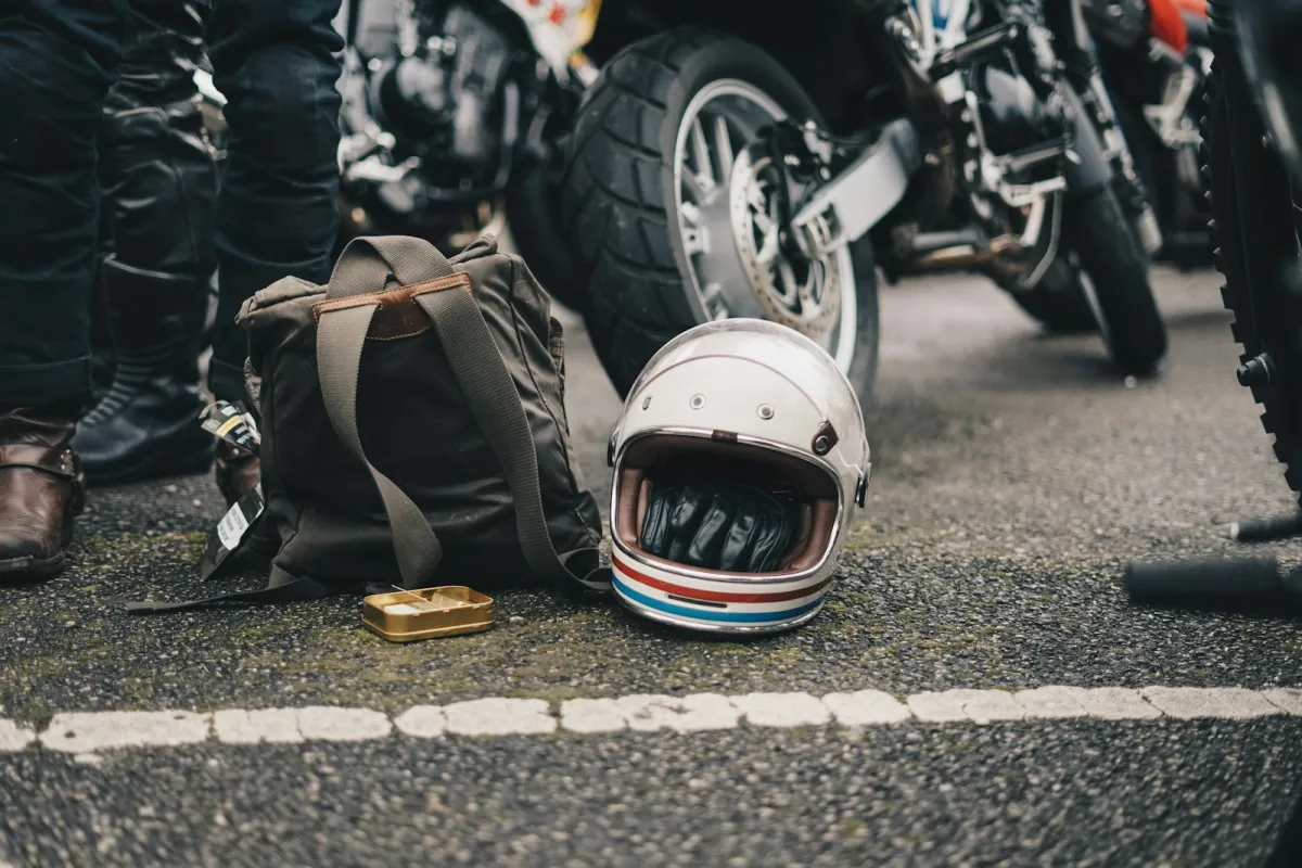 A white open-face motorcycle helmet and a canvas backpack on the tarmac beside parked motorbikes