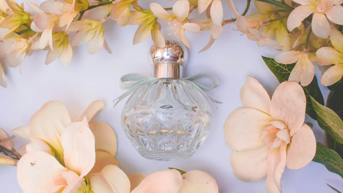 An unlabelled glass perfume bottle with a ribbon, surrounded by pale peach flowers An unlabelled glass perfume bottle with a ribbon, surrounded by pale peach flowers