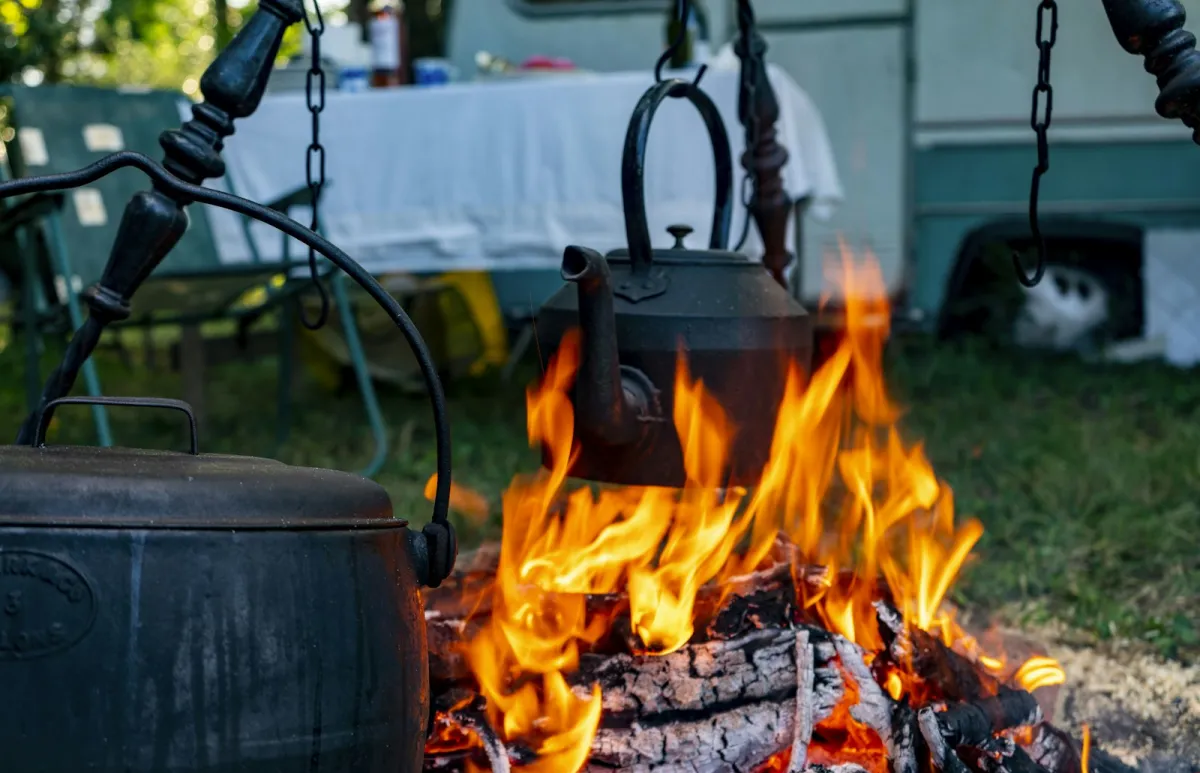 Cast iron pot and kettle hanging over a campfire beside a caravan