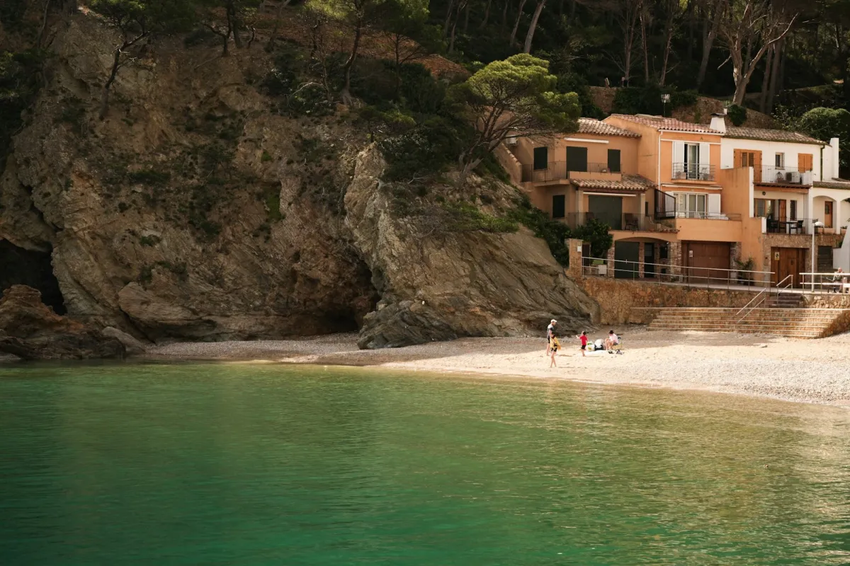 A small Costa Brava cove with green water and houses beneath a pine-covered cliff