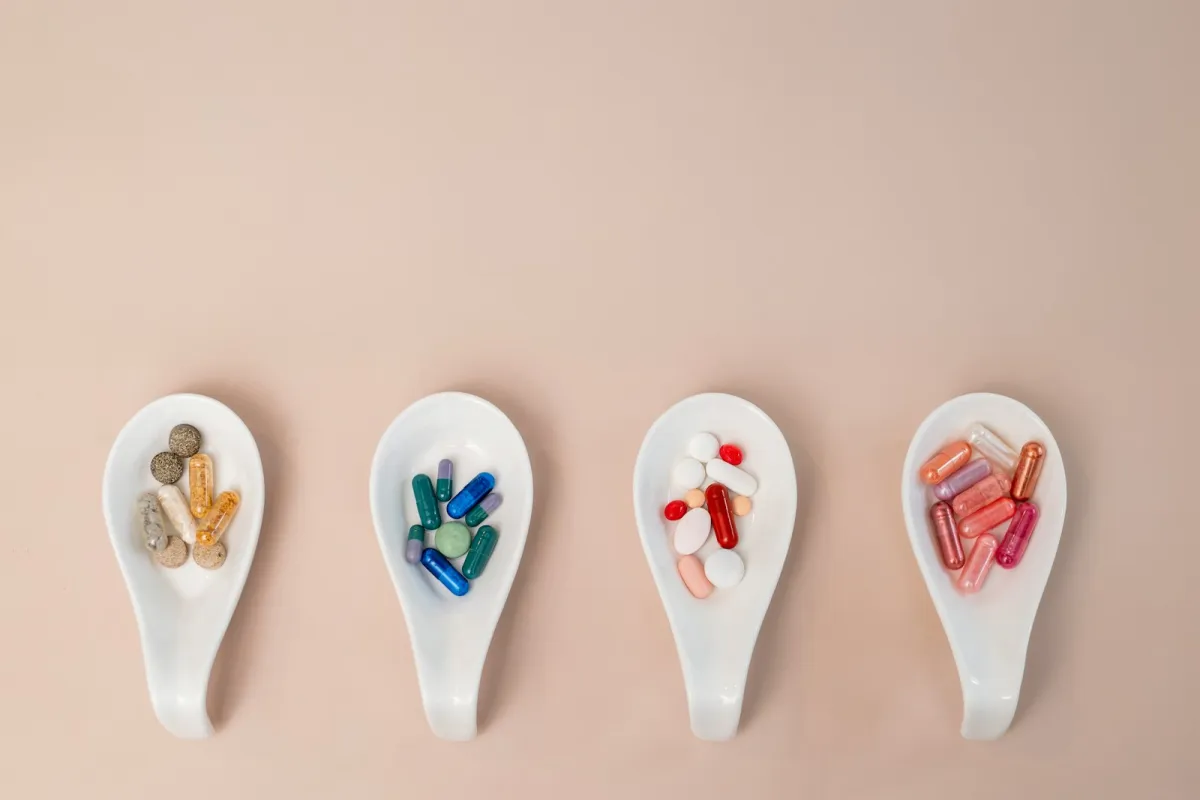 Four white ceramic spoons holding colourful capsules and tablets on a pale pink background