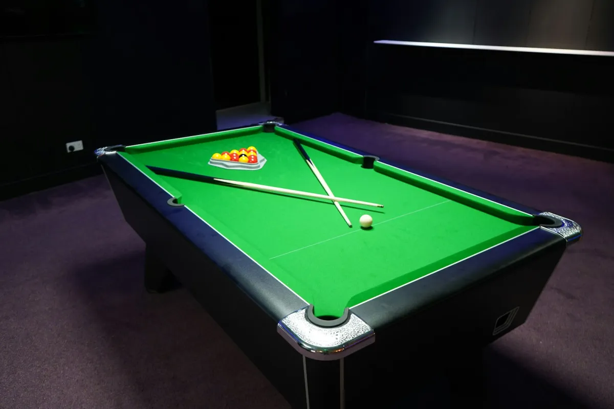 A green-baize pool table with racked balls and two crossed cues in a dark games room
