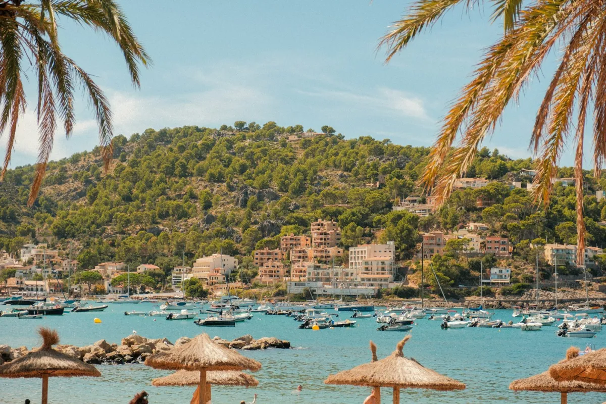 Boats anchored in a turquoise bay below wooded hills and hotels in Port de Sóller, Mallorca, framed by palm fronds Boats anchored in a turquoise bay below wooded hills and hotels in Port de Sóller, Mallorca, framed by palm fronds