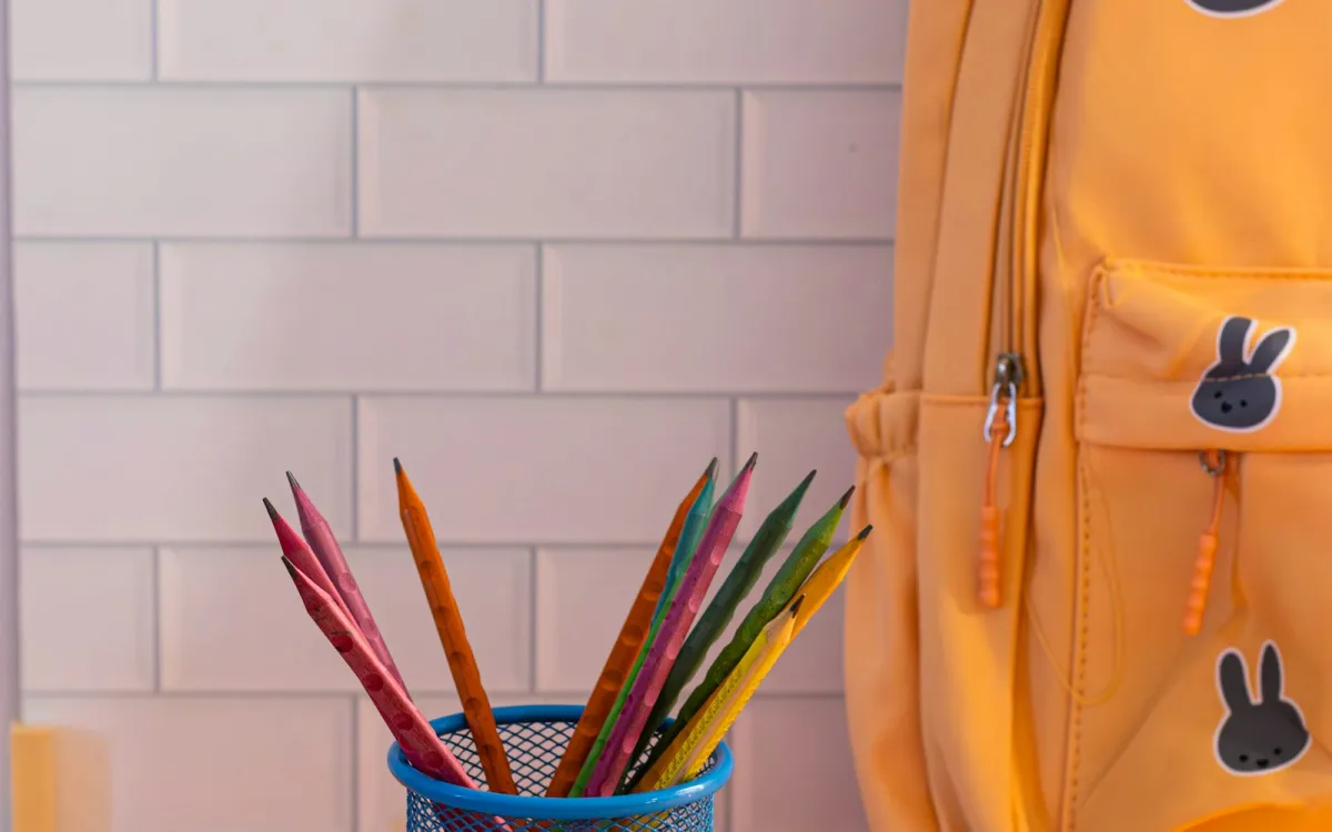 A yellow children's backpack beside a pot of coloured pencils A yellow children's backpack beside a pot of coloured pencils