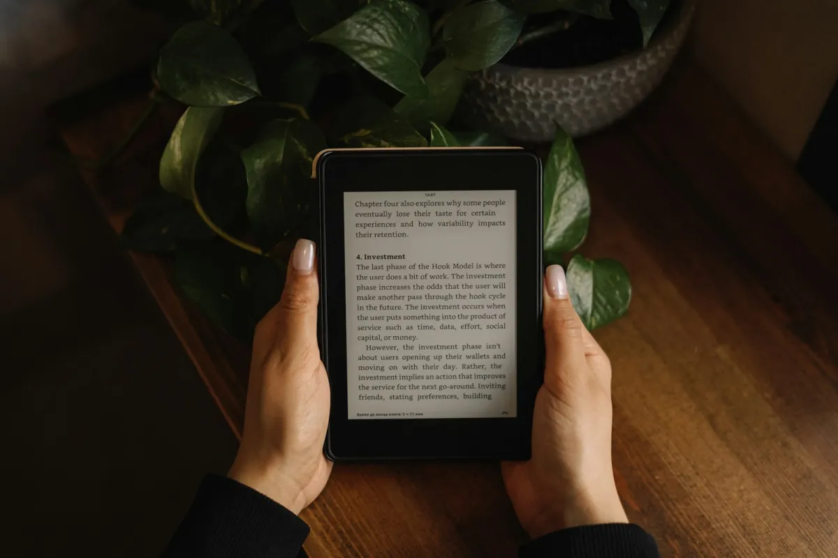 Two hands holding an e-reader showing a page of text above a wooden table and a trailing houseplant Two hands holding an e-reader showing a page of text above a wooden table and a trailing houseplant