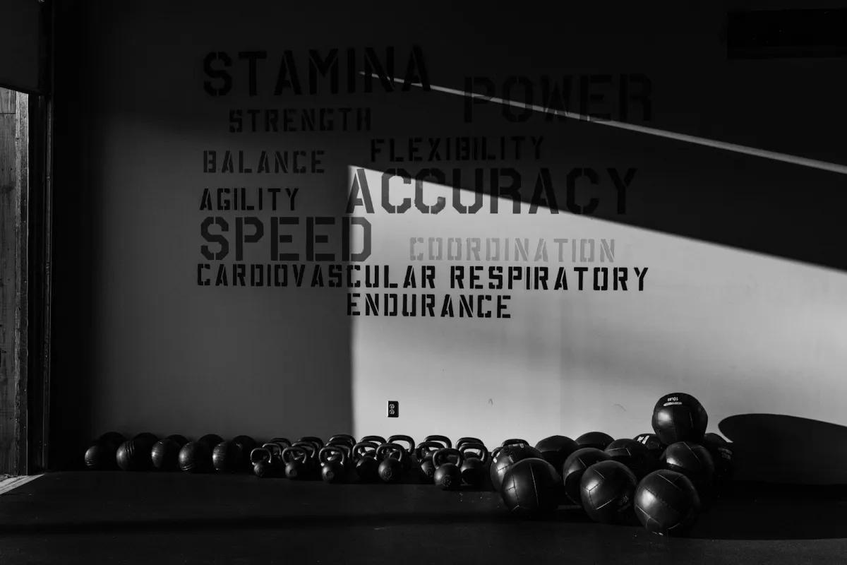 Black and white photo of kettlebells and medicine balls lined up on a gym floor beneath training words on the wall Black and white photo of kettlebells and medicine balls lined up on a gym floor beneath training words on the wall