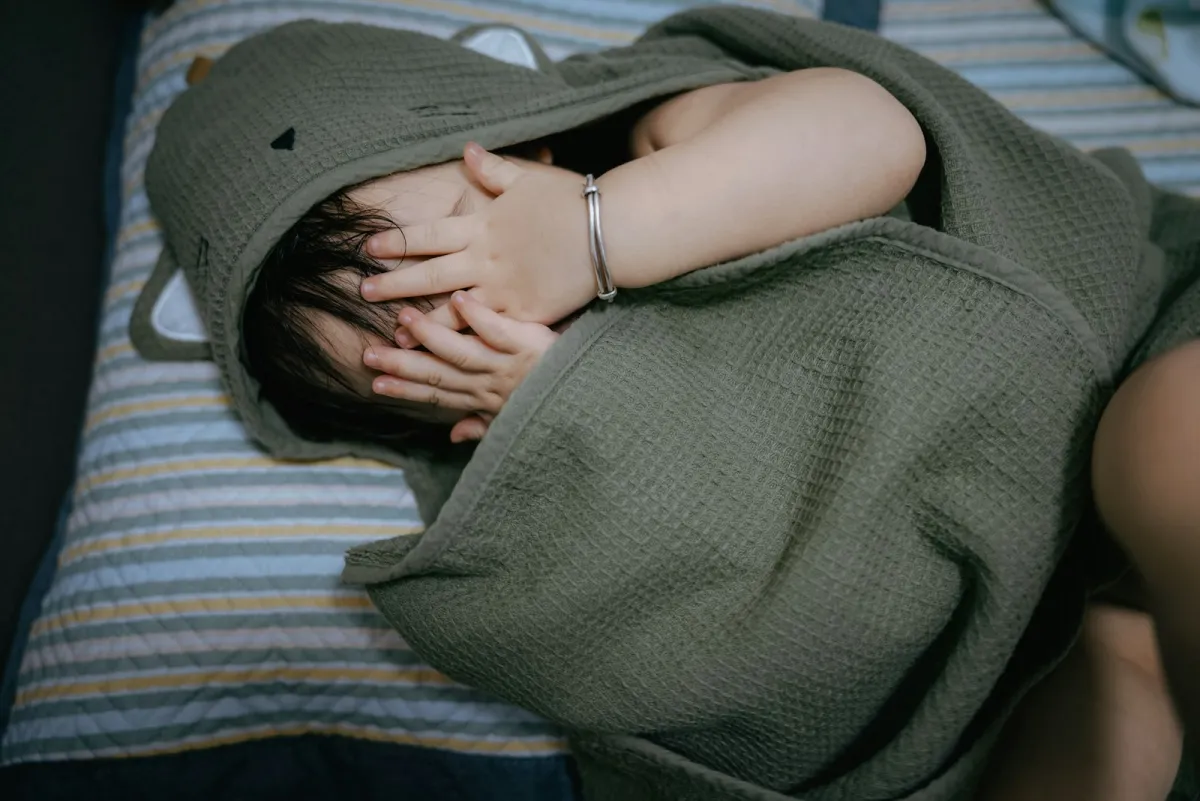 Young child wrapped in a green hooded towel, covering their face with their hands