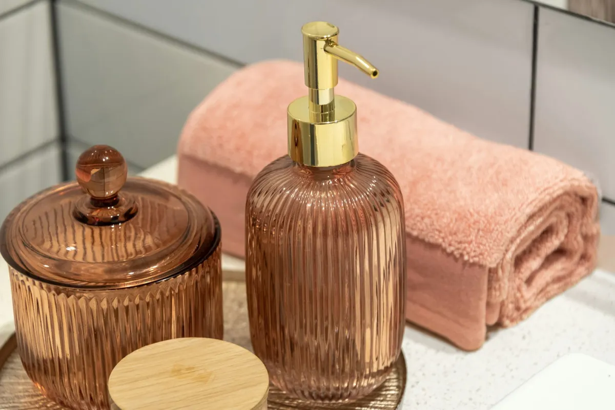 Ribbed glass soap dispenser with a gold pump beside a glass jar and a rolled pink towel