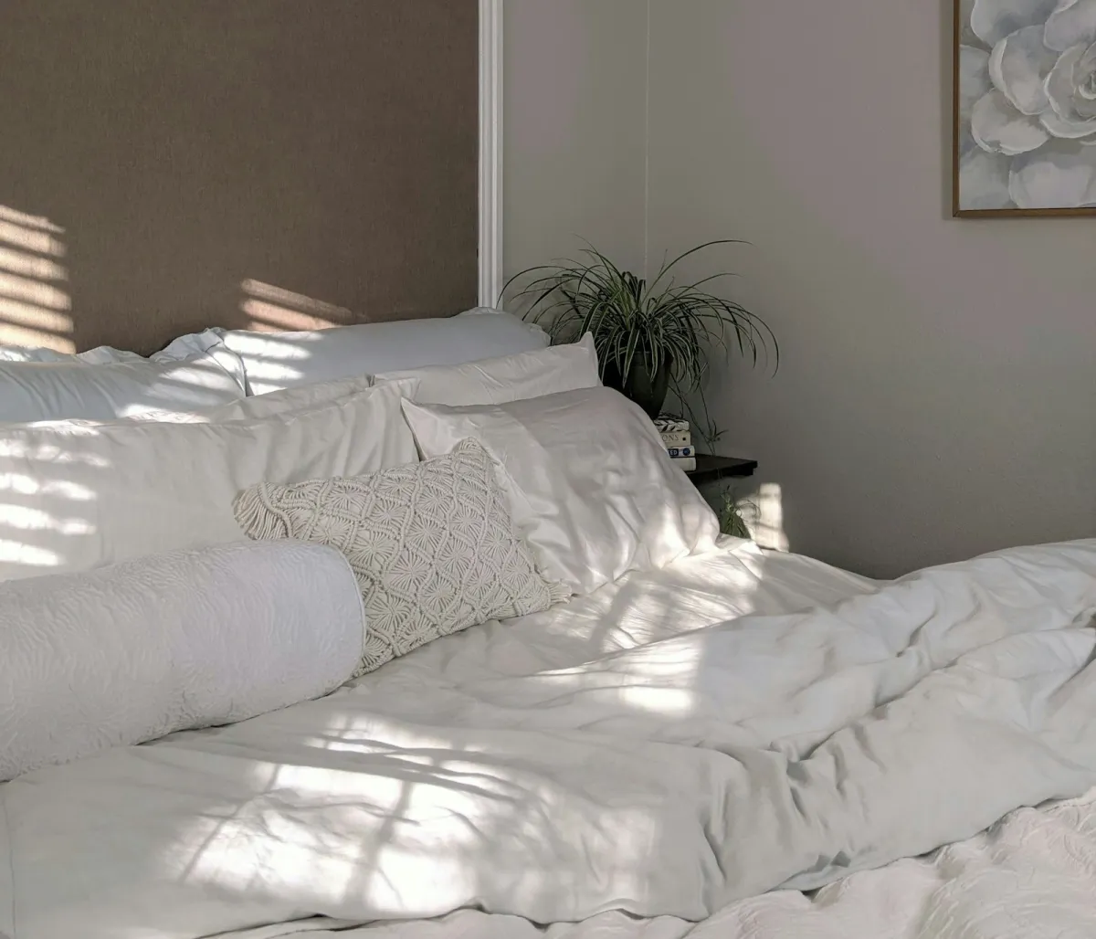 A made bed with white linen and cushions in a sunlit bedroom A made bed with white linen and cushions in a sunlit bedroom