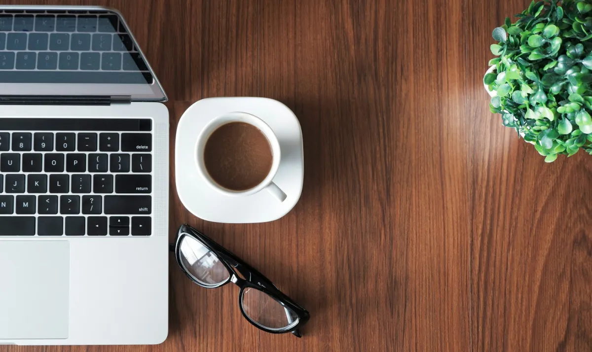 An open laptop, a cup of coffee, glasses and a potted plant on a wooden desk An open laptop, a cup of coffee, glasses and a potted plant on a wooden desk