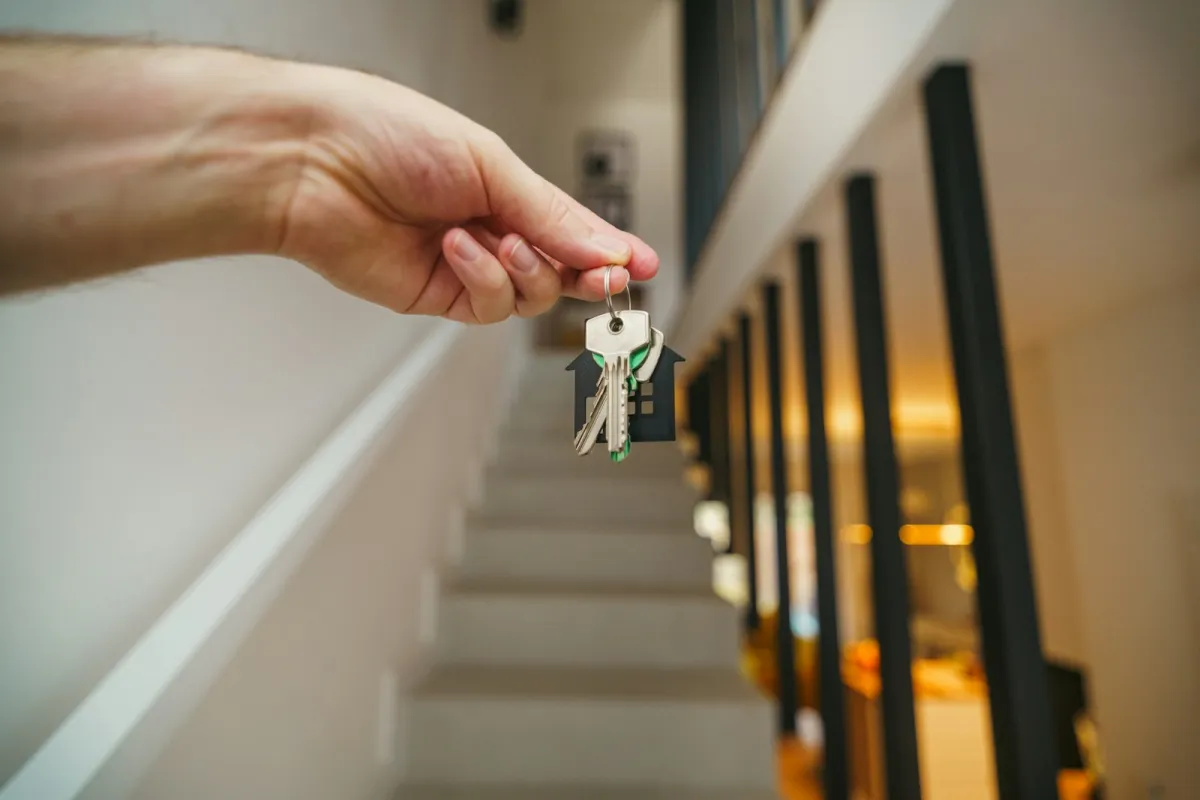 Hand holding a set of house keys on a house-shaped keyring in front of a staircase