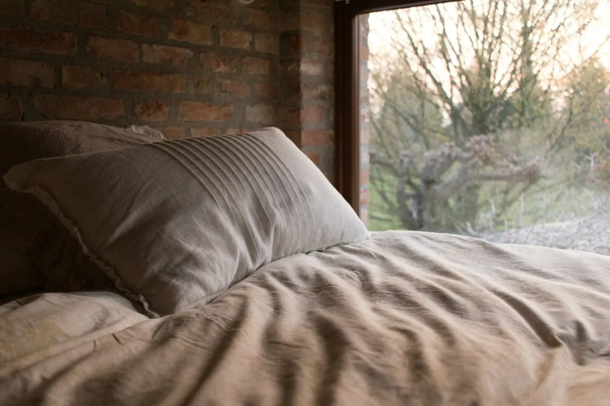 Pillow and rumpled linen duvet on a bed beside a large window