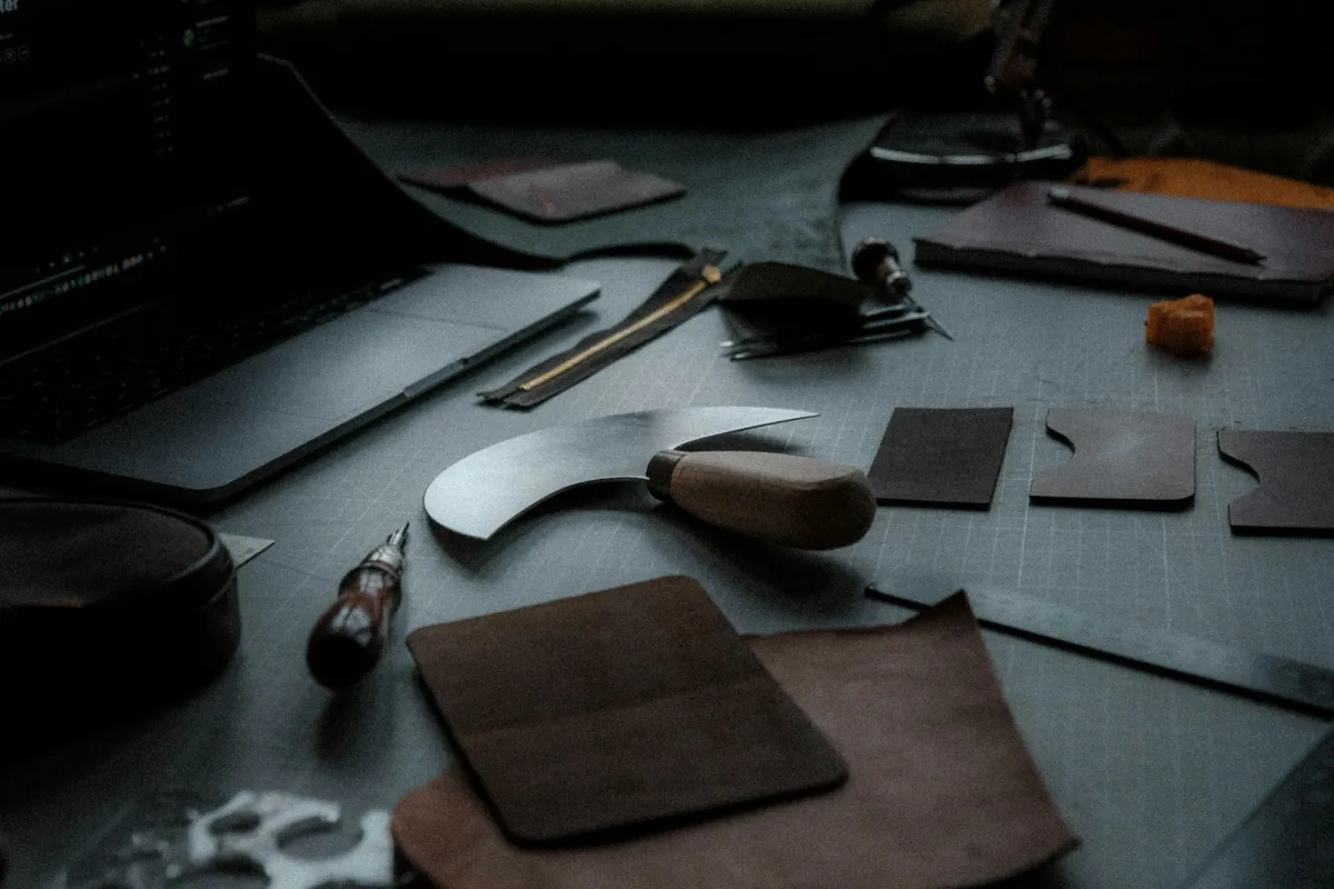 Leather-working tools, a curved knife and cut leather pieces on a dark workbench beside a laptop
