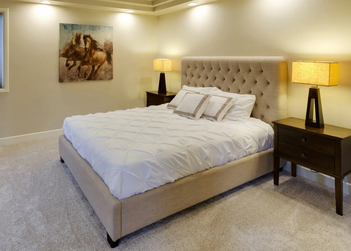 Double bed with a tufted beige headboard and white bedding between two lit bedside lamps