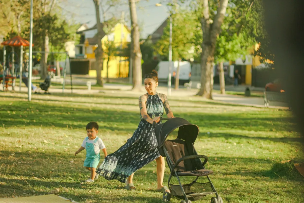 Woman pushing a pushchair through a sunny park with a toddler walking alongside