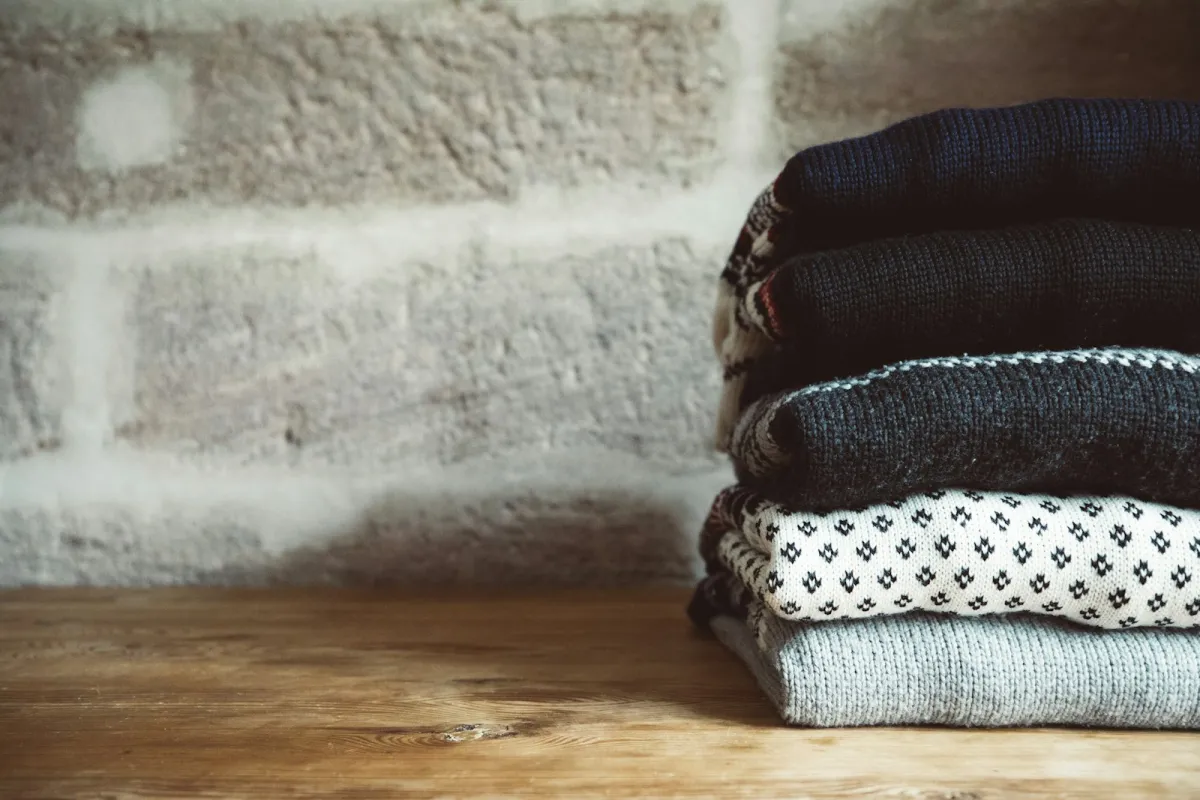 Stack of folded knitted jumpers on a wooden table against a brick wall