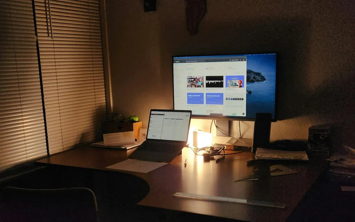 A laptop and a second monitor on a desk lit by a lamp in the evening A laptop and a second monitor on a desk lit by a lamp in the evening