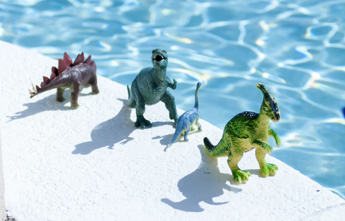 Four toy dinosaurs standing on the white edge of a sunny swimming pool